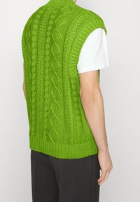 Pull sans manches en tricot vert avec motifs torsadés, ourlet côtelé et col montant, porté par-dessus un T-shirt blanc.