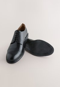 Chaussures à lacets en cuir noir avec une finition polie, design à bout droit, et semelle en caoutchouc noir texturée, exposées à côté de la semelle intérieure.