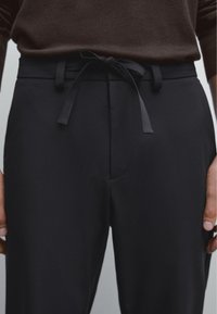 Pantaloni neri in tessuto leggero con vita a coulisse, texture liscia e vestibilità su misura, mostrati su uno sfondo neutro.