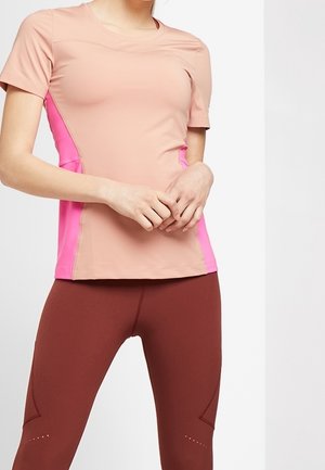 Maglietta sportiva rosa e pesca con maniche corte, texture liscia, design aderente e pannelli laterali a contrasto. Indossata con leggings marroni.