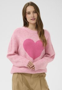 Femme aux longs cheveux bruns portant un pull rose avec un grand motif de cœur et un pantalon beige, les mains jointes à la taille.