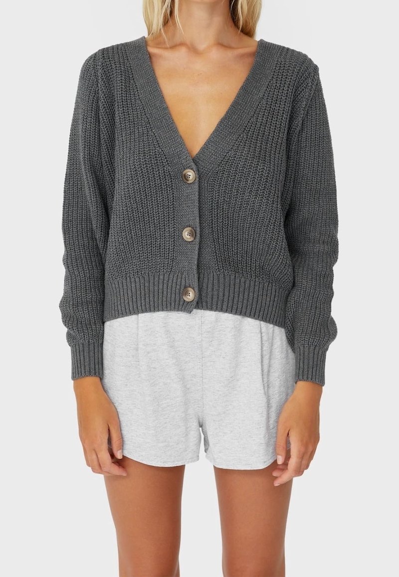 Woman wearing a charcoal gray chunky knit cardigan with three buttons over light gray shorts, standing against a plain background.