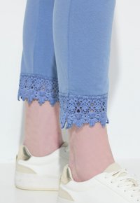 Ljusblå beskurna byxor med spetsdetaljer vid fållen, kombinerat med vita sneakers med metalliska accenter. Slät tygtextur.