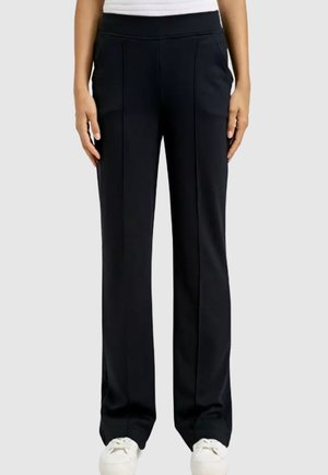 Woman wearing black straight-leg pants with a white elastic waistband and white sneakers, standing against a plain background.