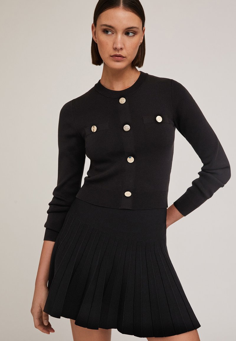 Cardigan en tricot noir avec un col rond, comportant six boutons dorés et deux petites poches. Associé à une mini-jupe plissée noire.