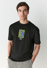 Black cotton t-shirt featuring a colorful shark graphic on the chest, framed by a bright green background with abstract lines.