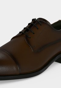 Chaussure habillée en cuir marron avec lacets noirs, bout arrondi et semelle basse noire sur fond clair.