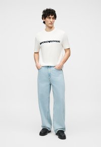 Νεαρός άνδρας με σγουρά μαλλιά που φοράει λευκό t-shirt Emporio Armani, ανοιχτόχρωμα τζιν με φαρδιά μπατζάκια και μαύρα παπούτσια, στέκεται με τα χέρια στις τσέπες.