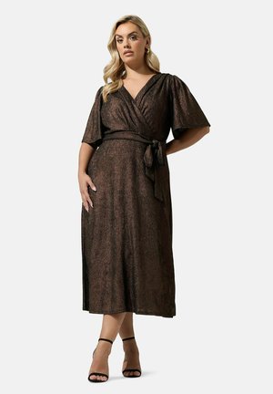 Femme portant une robe portefeuille marron scintillante avec des manches volantées, nouée à la taille, associée à des talons noirs à bride à la cheville, debout, une main sur la hanche.