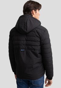 Giacca a vento nera con cappuccio, caratterizzata da sezioni trapuntate, polsini elastici e un logo prominente sul retro. Tessuto leggero e texturizzato.