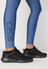 Μπλε κολάν με γυαλιστερή φινίρισμα και μαύρο λογότυπο "Casall"; συνδυασμένα με μαύρα αθλητικά παπο ύτσια από διχτυωτό ύφασμα με ενισχυμένη σόλα.