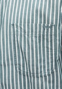 Ύφασμα με κάθετες ρίγες σε πράσινο και λευκό χρώμα, με ραμμένη ορθογώνια τσέπη που έχει μικρό κεντητό διπλό λογότυπο H στην πάνω δεξιά γωνία.