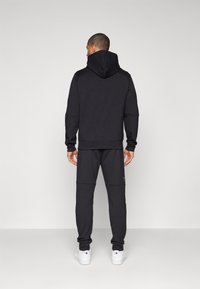 Svart hoodie med hög krage, kombinerad med matchande mjukisbyxor. Slät tyg, slim fit, och ribbade manschetter. Vita sneakers fulländar looken.
