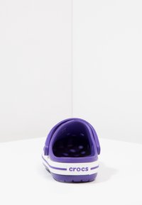 Zoccoli Crocs viola con cinturino posteriore, fori di ventilazione, strisce bianche e suola texturizzata. Vista posteriore che mostra il logo del marchio e un design arrotondato.