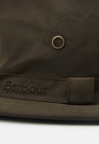 Brown hat made of water-resistant fabric, featuring a textured surface, an embossed brand name, and a metal vent detail.