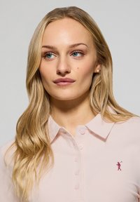 Ljusrosa polo-shirt med krage, med en knappslå med fem knappar och en liten broderad logotyp på vänster bröst. Slät textur.