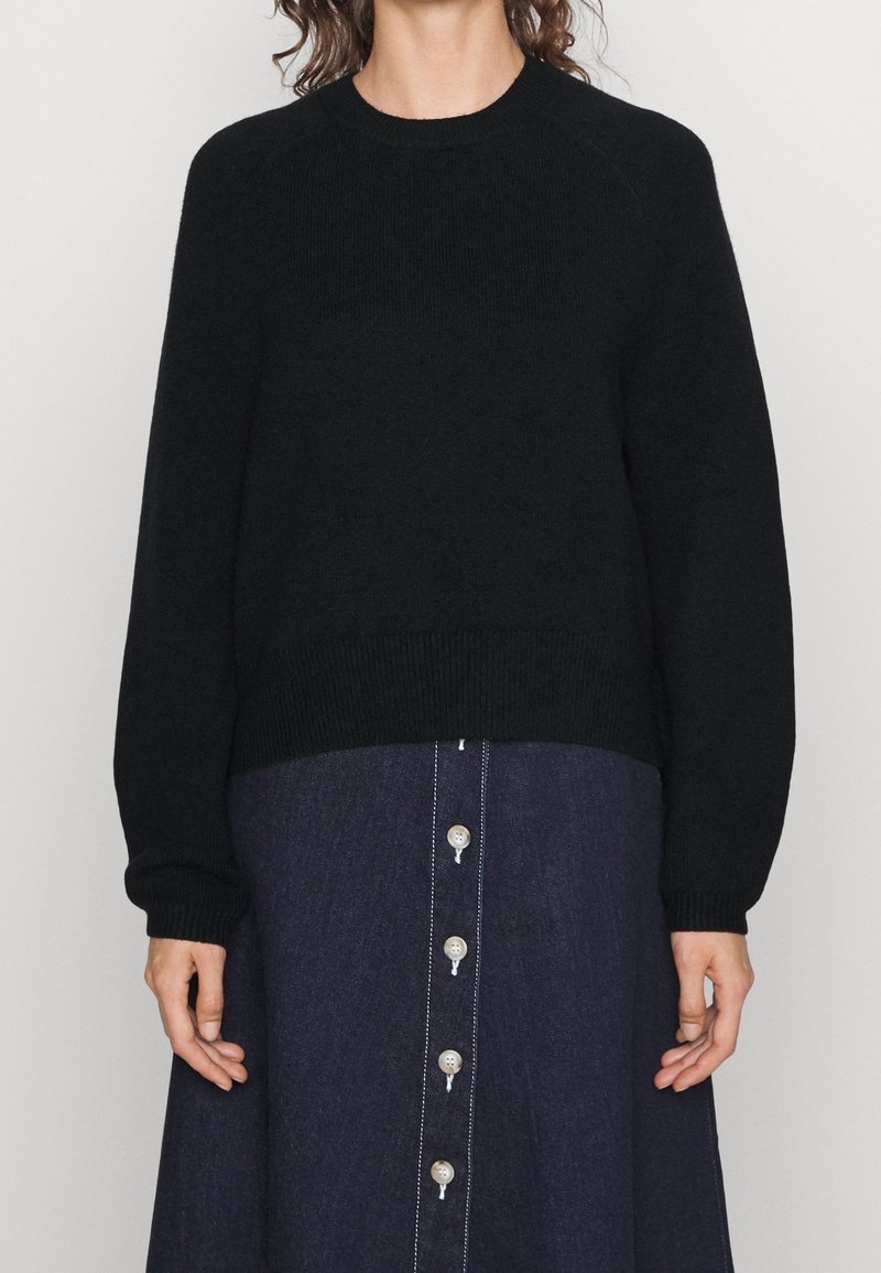 Femme portant un pull noir à manches longues et une jupe bleu foncé boutonnée, mains détendues le long du corps sur un fond neutre.