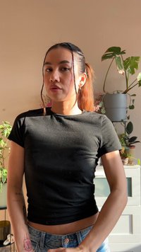 Schwarzes, eng anliegendes Kurzarmshirt und verwaschene hellblaue Jeans. Der Hintergrund zeigt grüne Pflanzen und eine hellfarbige Wand. Natürliches Licht.