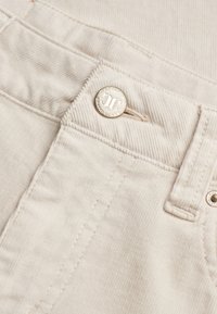 Nærbillede af beige fløjlsbukser med metaltrykknap og syningsdetaljer på linning og lommeområde.