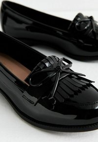 Mocassins en cuir verni noir avec des détails en frange et un accent en nœud. Texture lisse avec un bout arrondi et une semelle à profil bas.