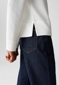 Persoon in een geweven witte langslijftop met een zijsplit, gecombineerd met donkerblauwe denimjeans, waarbij de achterzak zichtbaar is en de hand ontspannen naast het lichaam hangt.