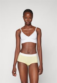 Bralette blanche avec des fines bretelles et un short en coton jaune avec une taille blanche imprimée "PIECES." Texture lisse, design ajusté.