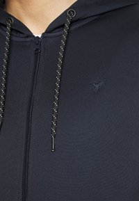 Marineblå hættetrøje med lynlås i tekstureret stof, der har et lille broderet logo og sorte snore med sølvdetaljer.