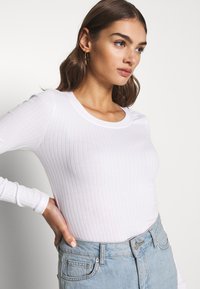 Weißes langärmeliges, gerippte Oberteil mit rundem Halsausschnitt, figurbetontem Schnitt und weicher Textur, kombiniert mit hochtaillierten hellblauen Jeans.