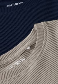 Nahaufnahme von gerippten Stoffkleidern in Beige und Marineblau, die Ausschnitte und teilweise den aufgedruckten Markennamen "TWO SOCI" zeigen.