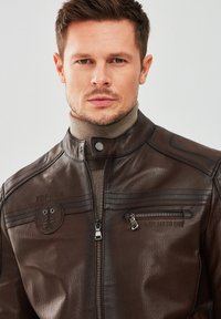 Brown leather jacket with zipper front, high collar, and embroidered details. Features side stripes and a zippered pocket on the chest.