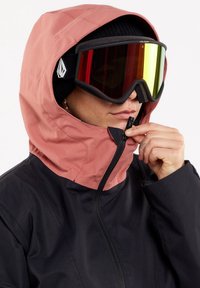Personne portant une veste à capuche noire et rose, un bonnet noir et des lunettes de ski miroir, remontant la fermeture éclair de la veste près du menton.