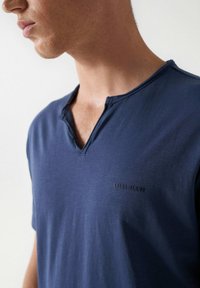 Tmavě modrá košile s krátkým rukávem a výstřihem do V, zdobená nenápadným logem "Salsa Jeans" na hrudi. Lehčí materiál, hladká textura.