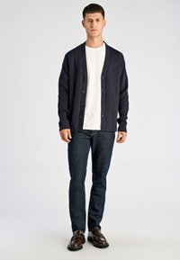 Granatowy prążkowany kardigan na pięć guzików, noszony na białej koszulce z okrągłym dekoltem. Stylizację dopełniają ciemnoniebieskie dżinsy i brązowe buty.