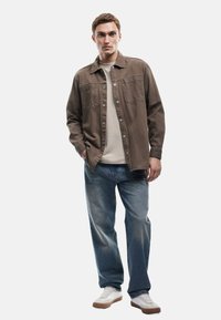 Camisa marrón de botones sobre una camiseta color beige, combinada con jeans azul claro y zapatillas blancas. El modelo está de pie contra un fondo liso.