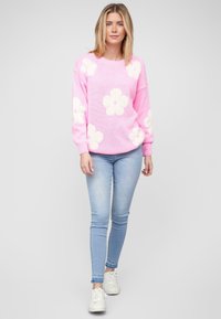 Roze gebreide trui met witte bloemenpatronen, ronde halslijn en een relaxte pasvorm. Gepaard met lichtblauwe skinny jeans en witte sneakers.