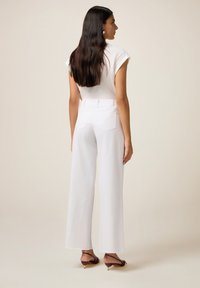 Femme aux longs cheveux noirs portant un pantalon large blanc taille haute, un top sans manches blanc et des chaussures à talons kitten brunes, de dos.