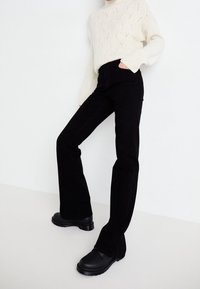 Jeans negros acampanados de mezclilla, combinados con un suéter tejido crema con un patrón de ondas. Calzado negro con suela gruesa.