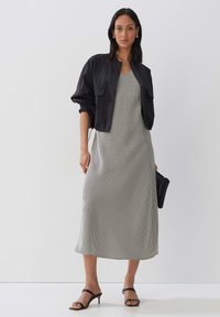 Schwarze Bomberjacke über einem ärmellosen, knielangen, karierten Kleid in Schwarz und Weiß. Eine schwarze Clutch und hochhackige Sandalen runden den Look ab.