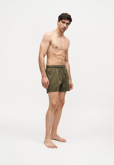 Νέος άνδρας με γυμνασμένο σώμα, που φοράει χακί πράσινα μαγιό, όρθιος ξυπόλητος σε λευκό φόντο.