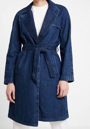 Vrouw draagt een donkerblauwe denim trenchcoat met strikceintuur, kraag met inkeping en zijzakken over een wit top en broek.
