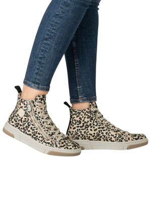 Personne portant des baskets montantes à imprimé léopard avec fermetures éclair latérales et un jean skinny bleu, debout avec les pieds croisés.