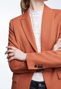 Blazer in misto lana arancione con rever a lancia e tre polsini con bottone nero, indossato sopra un top in pizzo bianco. Focalizzato su vestibilità su misura e texture.