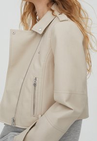 Beige Kunstlederjacke mit einem kurzen Design, diagonalem Reißverschluss und silbernen Reißverschlüssen an den Seitentaschen. Mit einem Reverskragen.