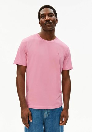 Mann trägt ein schlichtes pinkfarbenes T-Shirt mit kurzen Ärmeln und blaue Jeans, steht mit neutralem Ausdruck vor weißem Hintergrund.