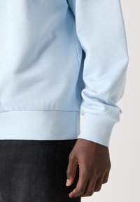 Sudadera azul claro con puños y dobladillo acanalados. Presenta una textura suave y un corte relajado, exhibida sobre un fondo de tela negra.