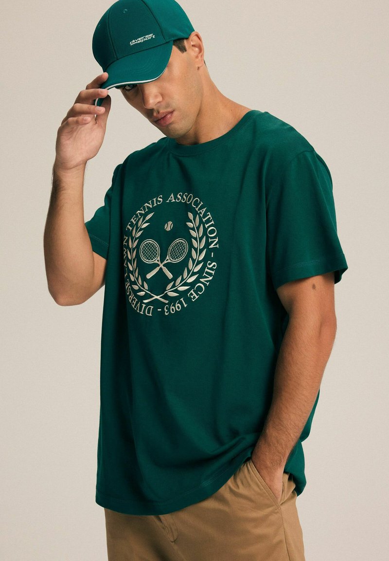 Tealowa bawełniana koszulka z okrągłym dekoltem z nadrukiem tematycznym związanym z tenisem i laurowymi gałązkami. Dopasowana tealowa czapka uzupełnia strój.