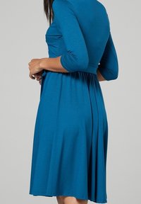 Tyrkysov�é šaty ke kolenům vyrobené z měkké látky, s třičtvrtinovými rukávy a přiléhavým živůtkem, které se na pase rozšiřují.