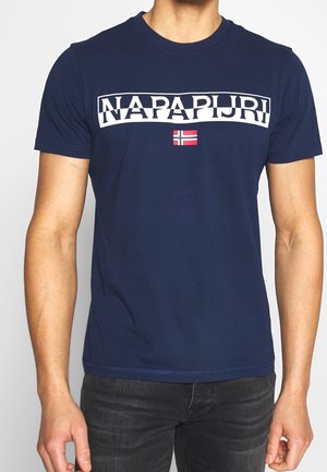 Mężczyzna noszący granatową koszulkę Napapijri z białym logo i małą norweską flagą na środku, połączoną z ciemnymi jeansami.