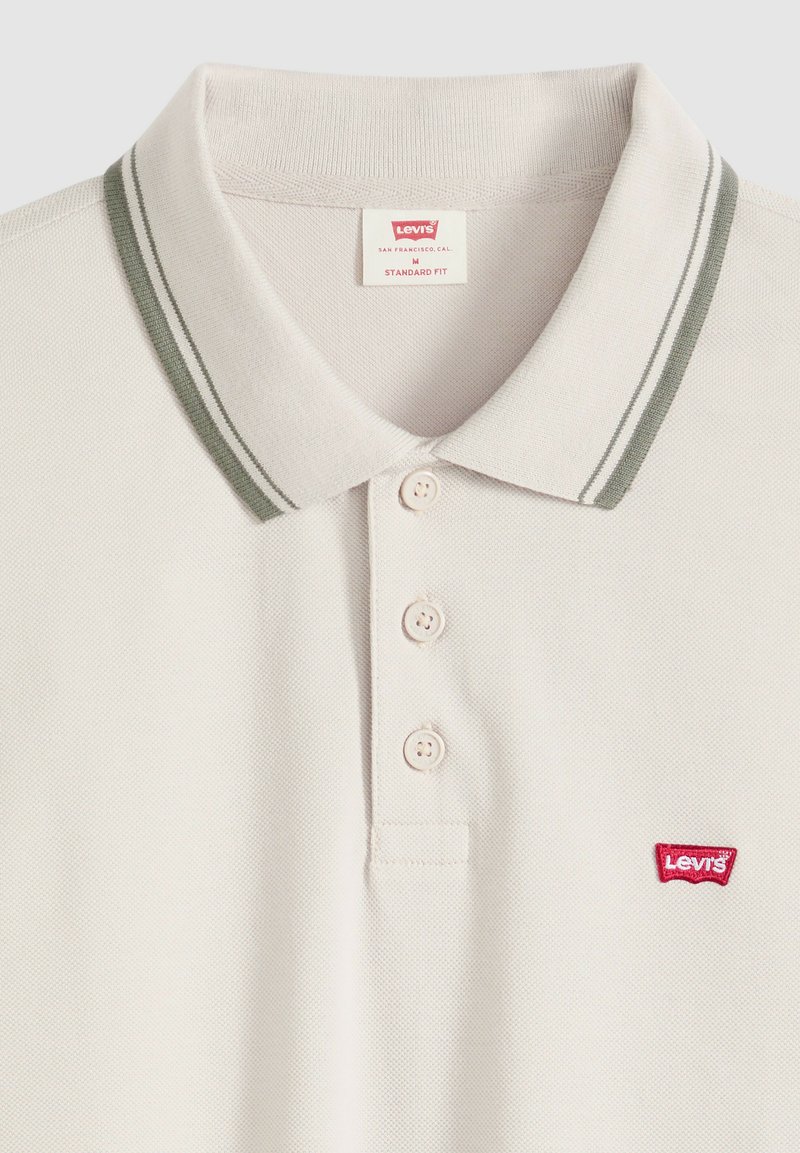 Polo Levi's de couleur crème avec trois boutons, double rayures grises sur le col, et un écusson logo Levi's rouge sur la poitrine.