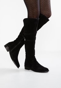 Botas de ante negras hasta la rodilla con tacón plano, que presentan una cremallera lateral y un detalle de correa en contraste alrededor de la pantorrilla. Acabado texturizado visible.
